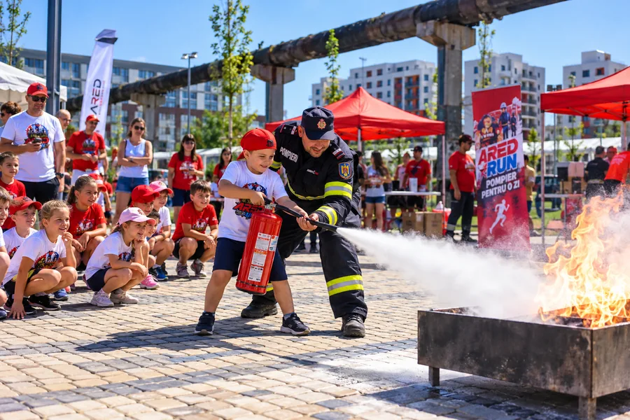 Un copil folosește un stingător (extinctor) pentru a stinge un foc de exercițiu, ajutat de un pompier